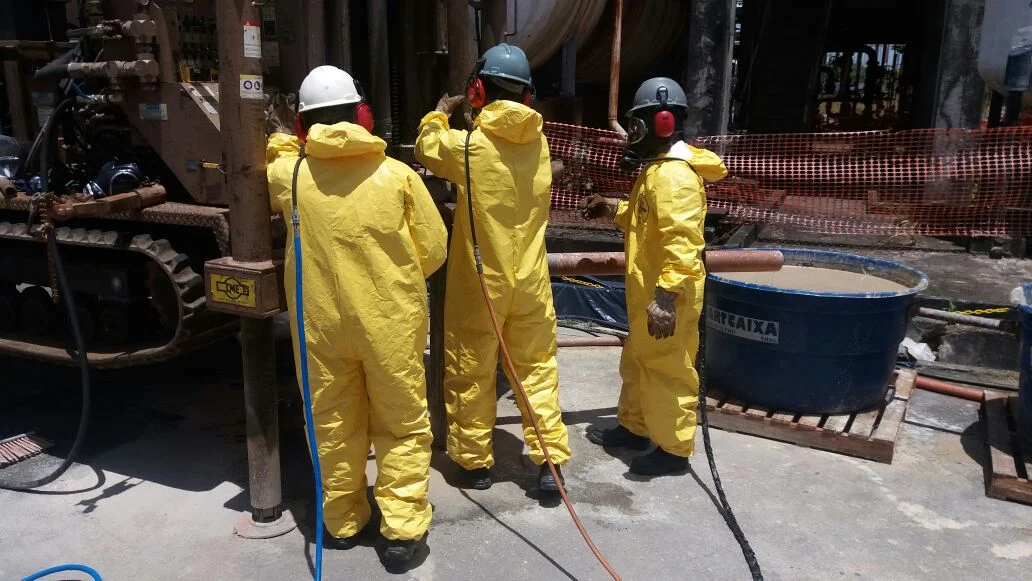 Perfuração de poço de monitoramento em área contaminada, com equipe em EPI de proteção química.